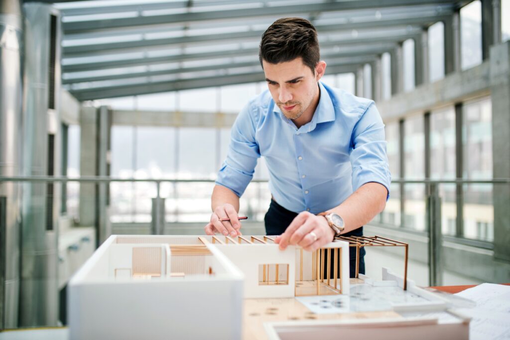 Junger Geschäftsmann oder Architekt mit einem Modell eines Hauses, der im Büro steht und arbeitet.